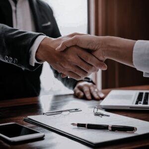 Two professionals shaking hands over a desk with documents and a laptop, symbolizing a business agreement.